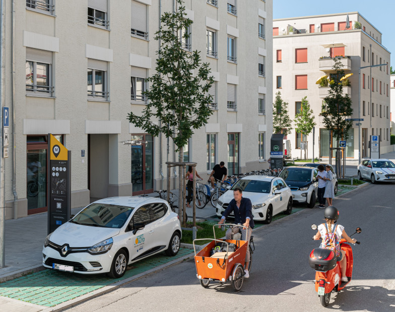 Das Foto zeigt ein Wohnhaus mit einer vorgelagerten Mobilitätstation mit Fahrrädern, Lastenrädern, Autos und Motorrollern