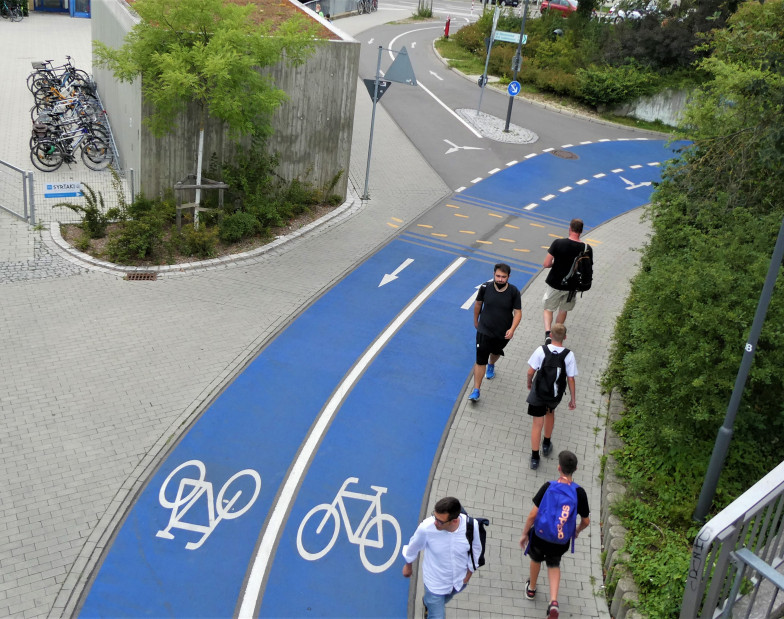 Beidseitiger Fahrradweg mit blauer Fahrbahn von oben. Daneben laufen Fußgänger*innen.