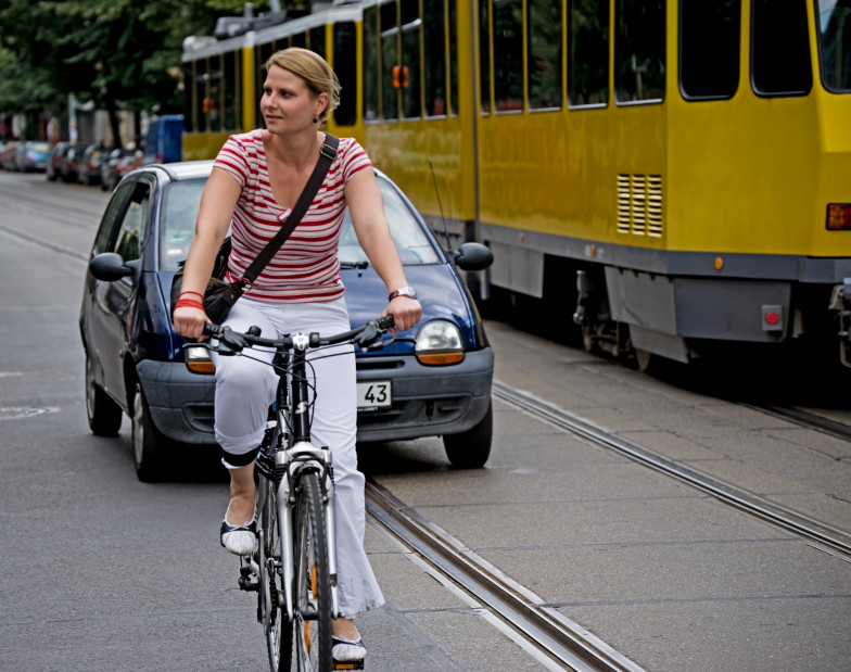 Foto einer Radfahrerin im Straßenverkehr, im Hintergrund Auto und Tram