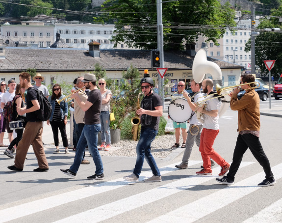 Foto: die Mitglieder einer Brass-Band laufen in einer Reihe hintereinander über einen Fußgängerüberweg