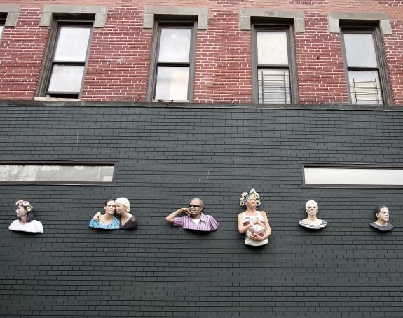 Foto: an einer dunklen Hauswand sind die Skulpturen angebracht, die unterschiedliche Menschen darstellen