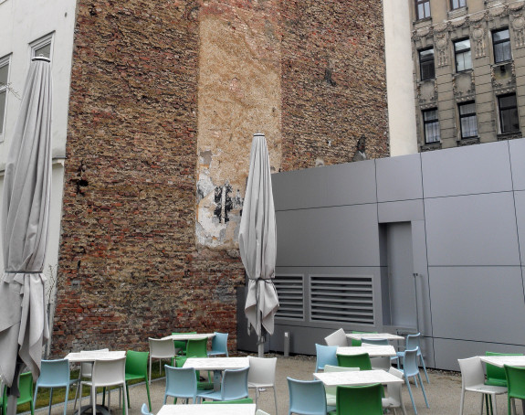 Foto: Baulücke zwischen Altbauten, auf der sich ein Café mit Tischen. Stühlen und Sonnenschirmen befindet.