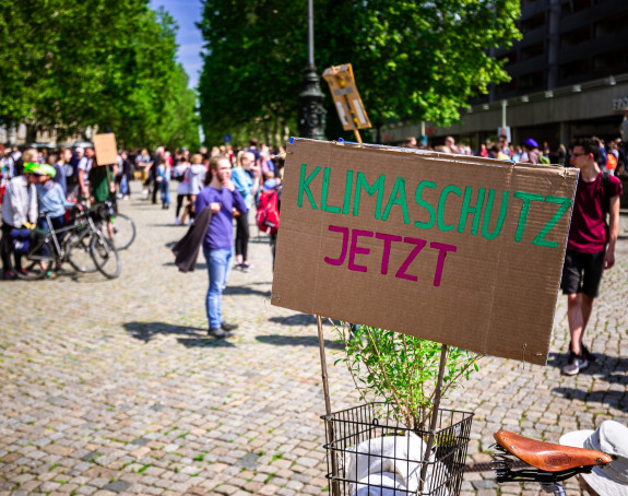 Foto: Schild mit Aufschrift "Klimaschutz jetzt", Demonstration im Hintergrund 