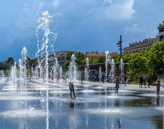 Das Bild zeigt ein Wasserspiel in einer Stadt, in dem sich Menschen abkühlen.