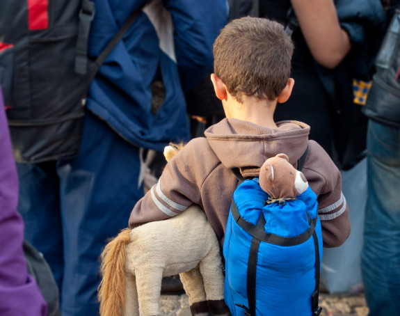 Gruppen wartender Menschen, im Vordergrund Kind mit Rucksack und Teddy von hinten