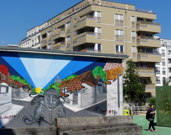 Foto eines Mehrfamilienhauses, davor stehend ein niedriges Gebäude mit Graffiti