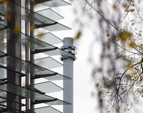 Foto: Schornstein eines Energieversorgers zwischen einem Neubau und einem Baum